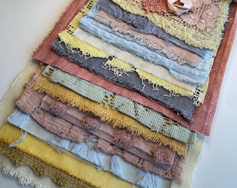 May include: A stack of rectangular fabric swatches in various colors including yellow, blue, pink, and mauve. The pieces feature frayed edges, lace textures, and woven patterns. A light pink ribbon is tied in a bow at the top of the fabric collection.