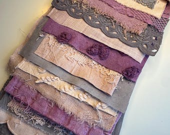 May include: A collection of layered fabric strips in shades of purple, gray, and beige. The strips feature varied textures including frayed edges, scalloped borders, intricate lace, embroidered floral knots, and a tied ribbon at the top.