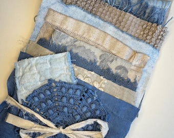 May include: A collection of layered fabric scraps in shades of blue and beige, featuring various textures including frayed edges, woven patterns, embroidered floral details, and a circular blue crocheted piece tied with a white fabric ribbon.