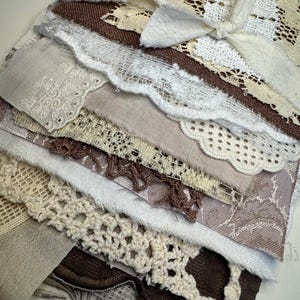 Puede incluir: Un collage textil en capas que presenta varios retazos de tela en tonos marrón, crema y blanco. La composición incluye piezas de encaje, lino tejido, algodón texturizado y adornos bordados, todo asegurado con un lazo de tela central.