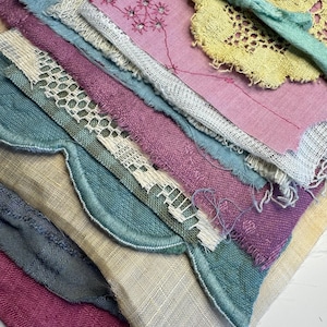 May include: A stack of layered fabric scraps featuring various textures and colors including yellow, teal, pink, and purple. The collection includes pieces of lace, embroidered linen, and frayed edges, with a light green ribbon tied around the top layer.