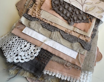 May include: A collection of layered fabric swatches in neutral tones including beige, cream, brown, and white. The stack features various textures such as crochet lace, tufted fabric, frayed linen, and a floral patterned paper wrap tied with a ribbon.