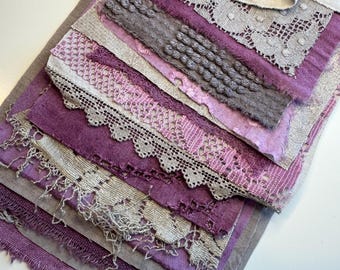 May include: A layered textile composition featuring various shades of purple and grey fabric scraps. The piece includes textured linen, intricate beige lace, embroidered floral patterns, and frayed edges, all secured with a light grey fabric ribbon tie.