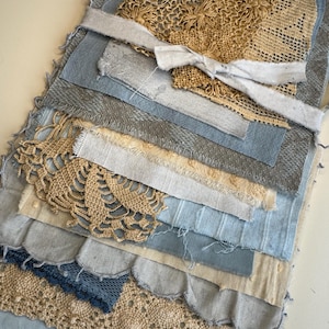 Puede incluir: Una obra de arte textil en capas con telas en tonos azules, beige y grises. Incluye encaje, materiales tejidos y una cinta gris claro atada con un lazo. El diseño tiene un aspecto rústico y artesanal, con diversas texturas.