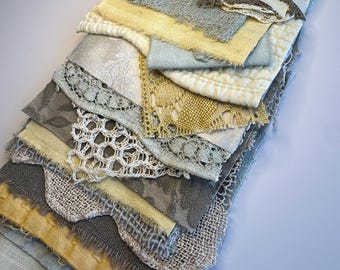 May include: A collection of layered fabric scraps in shades of yellow, gray, beige, and light blue. The assortment includes various textures such as woven linen, quilted cotton, delicate lace, and embroidered mesh, all stacked in a vertical arrangement.