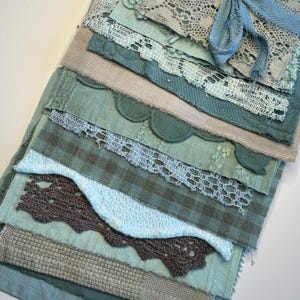 May include: A selection of fabric swatches in shades of blue, green, and beige, bound with a blue ribbon. The swatches feature lace, textured weaves, and patterned fabrics, creating a layered and tactile display of textiles.