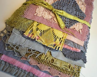 May include: A layered textile collage featuring various fabric scraps, including frayed linen, intricate lace, and crocheted elements in shades of dusty rose, sage green, yellow, and grey, all secured together with a thin, tied green ribbon.