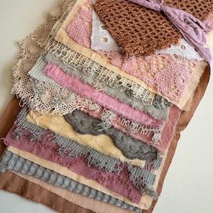 May include: A collection of fabric swatches in various shades of pink, brown, and gray, with lace and textured details. The swatches are tied together with a mauve ribbon. The textiles include crochet and fringed edges, creating a layered effect.