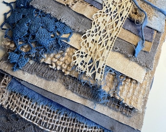 May include: A collection of layered fabric scraps in shades of blue, beige, and gray, featuring various textures including woven linen, coarse burlap, and delicate crochet. A strip of cream-colored lace and a blue string tie the pieces together.