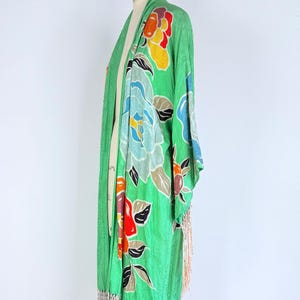 Puede incluir: Bata verde estilo kimono con un estampado floral en tonos azules, rojos, amarillos y naranjas. La bata tiene mangas largas y un ribete de flecos color melocot&oacute;n. La prenda se exhibe en un maniqu&iacute;.