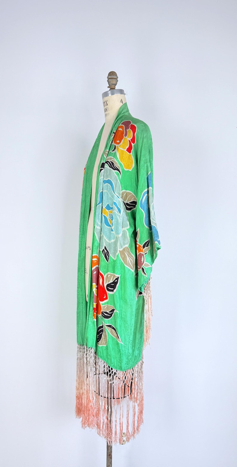 Puede incluir: Bata verde estilo kimono con un estampado floral en tonos azules, rojos, amarillos y naranjas. La bata tiene mangas largas y un ribete de flecos color melocot&oacute;n. La prenda se exhibe en un maniqu&iacute;.