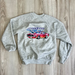 Puede incluir: Una sudadera gris jaspeada con un gráfico de un coche Mustang vintage rojo y naranja y la palabra "mustang" en azul y blanco. La sudadera de cuello redondo y manga larga está hecha de un material suave.