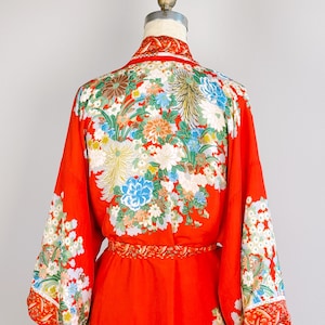 May include: A vibrant red kimono featuring an intricate floral pattern with chrysanthemums and other blossoms in shades of white, blue, and green. The garment includes wide sleeves, a matching waist sash, and a detailed collar design.