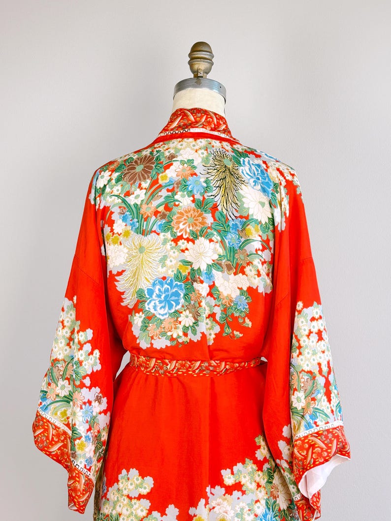 May include: A vibrant red kimono featuring an intricate floral pattern with chrysanthemums and other blossoms in shades of white, blue, and green. The garment includes wide sleeves, a matching waist sash, and a detailed collar design.