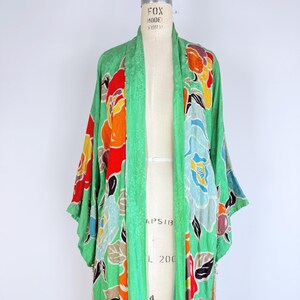 Puede incluir: Un kimono verde vintage con un estampado floral en rojo, naranja, amarillo y azul. El kimono se exhibe en un maniqu&iacute;. Las mangas son largas y la tela tiene una textura sutil. El kimono tiene un detalle de flecos en la parte inferior.