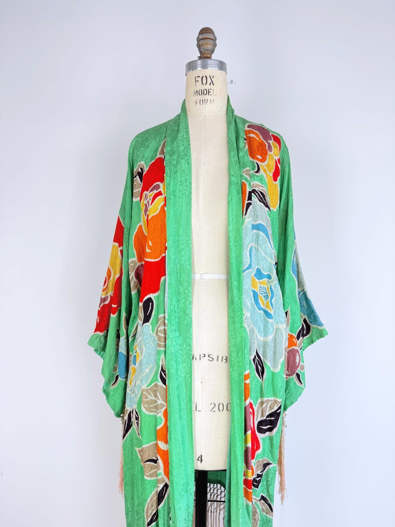Puede incluir: Un kimono verde vintage con un estampado floral en rojo, naranja, amarillo y azul. El kimono se exhibe en un maniqu&iacute;. Las mangas son largas y la tela tiene una textura sutil. El kimono tiene un detalle de flecos en la parte inferior.