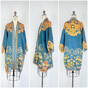 May include: A vintage blue kimono with an open front, displayed on a mannequin. The kimono features intricate floral and geometric patterns in shades of orange, yellow, and blue. The sleeves are wide, and the garment has a flowing silhouette.