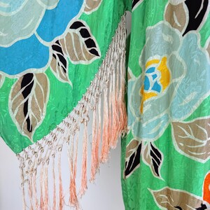 Puede incluir: Una prenda tipo kimono verde vibrante con un gran estampado floral en tonos azules, amarillos y rojos. La tela tiene una textura sutil y est&aacute; adornada con flecos color crema con acentos melocot&oacute;n. El dise&ntilde;o floral incluye hojas negras y marrones.
