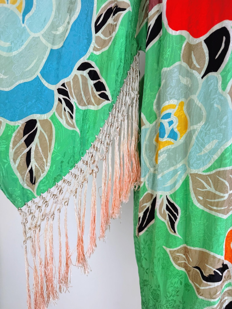 Puede incluir: Una prenda tipo kimono verde vibrante con un gran estampado floral en tonos azules, amarillos y rojos. La tela tiene una textura sutil y est&aacute; adornada con flecos color crema con acentos melocot&oacute;n. El dise&ntilde;o floral incluye hojas negras y marrones.