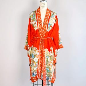 May include: A bright orange kimono robe featuring a detailed floral pattern in shades of white, blue, and green. The garment has wide sleeves, a matching tie belt at the waist, and a decorative border along the front opening and sleeve edges.