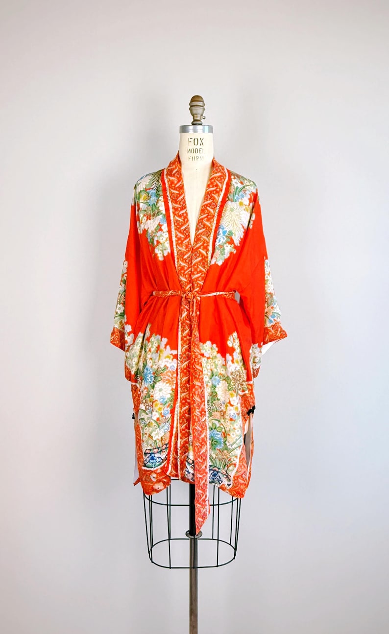 May include: A bright orange kimono robe featuring a detailed floral pattern in shades of white, blue, and green. The garment has wide sleeves, a matching tie belt at the waist, and a decorative border along the front opening and sleeve edges.