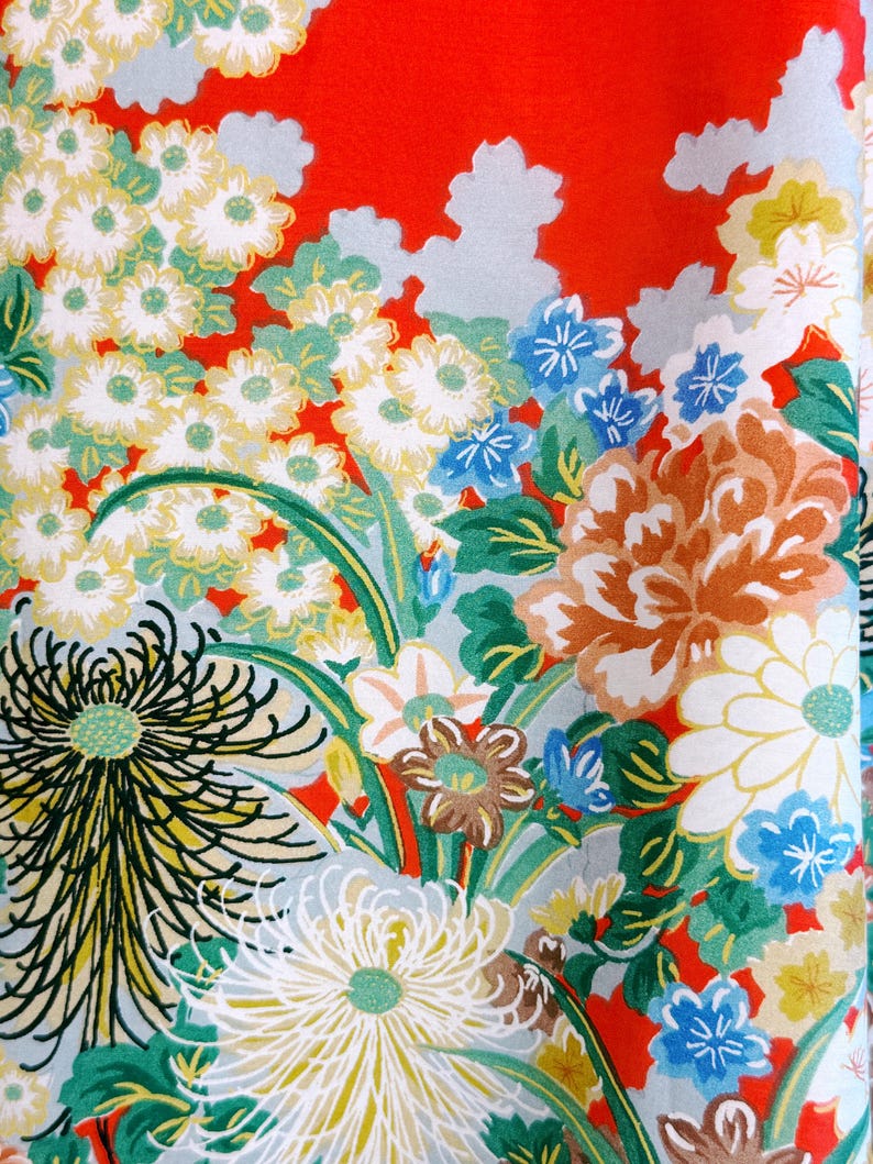 May include: A vibrant red fabric featuring a dense floral pattern with white, yellow, and blue chrysanthemums, green leaves, and light gray cloud-like shapes. The design includes large, detailed flower heads with thin petals and smaller blossoms.