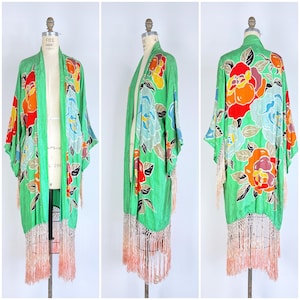Puede incluir: Una bata estilo kimono verde con un estampado floral en rojo, naranja, azul y amarillo. La bata tiene mangas largas y un ribete de flecos en color melocot&oacute;n claro. La prenda se exhibe en un maniqu&iacute;.