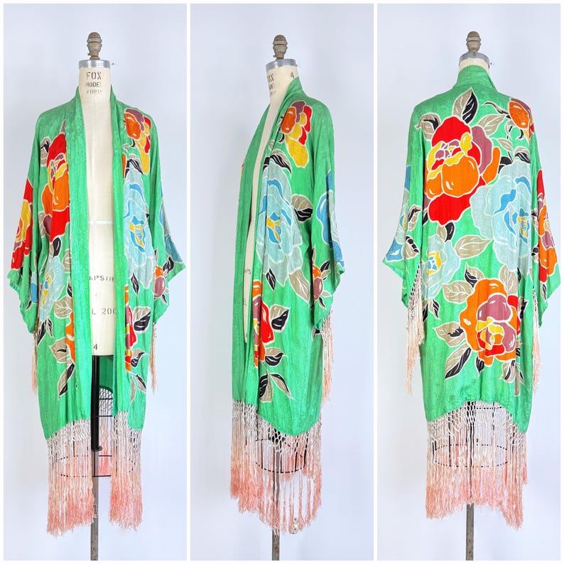 Puede incluir: Una bata estilo kimono verde con un estampado floral en rojo, naranja, azul y amarillo. La bata tiene mangas largas y un ribete de flecos en color melocot&oacute;n claro. La prenda se exhibe en un maniqu&iacute;.