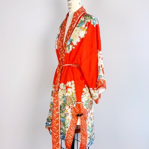 May include: A vibrant orange kimono robe featuring a detailed floral pattern with white, yellow, and blue blossoms. The garment includes a matching tie belt, wide sleeves, and a decorative border along the front opening and hemline.