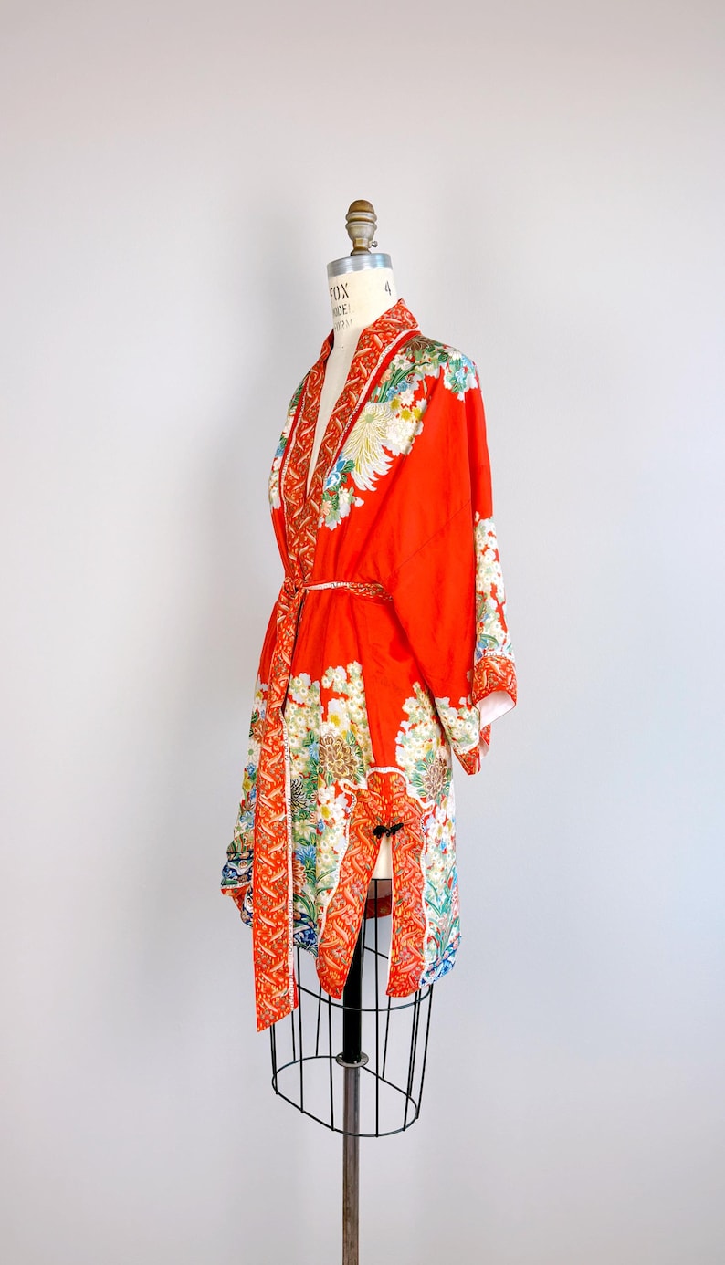 May include: A vibrant orange kimono robe featuring a detailed floral pattern with white, yellow, and blue blossoms. The garment includes a matching tie belt, wide sleeves, and a decorative border along the front opening and hemline.