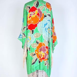 Puede incluir: Una bata verde estilo kimono con un estampado floral en rojo, naranja y azul. La bata tiene un ribete de flecos en color melocot&oacute;n claro. La prenda se exhibe en un maniqu&iacute; sobre un fondo blanco.