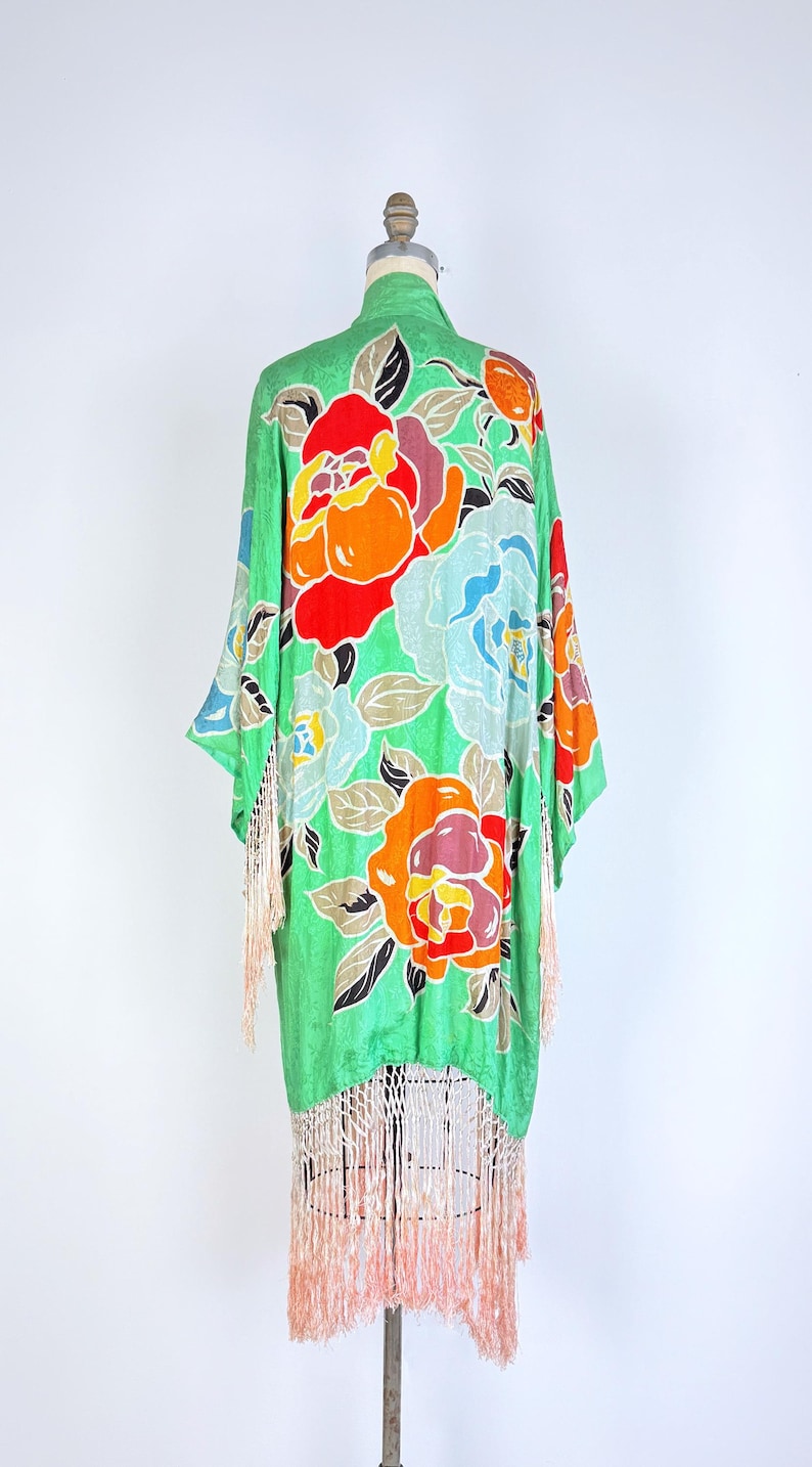 Puede incluir: Una bata verde estilo kimono con un estampado floral en rojo, naranja y azul. La bata tiene un ribete de flecos en color melocot&oacute;n claro. La prenda se exhibe en un maniqu&iacute; sobre un fondo blanco.