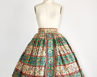 Puede incluir: Una falda vintage completa con estampado floral en tonos verdes, beige y rojos. La falda tiene una cintura fruncida y una silueta completa y fluida.