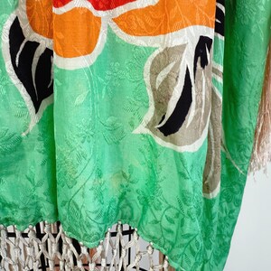Puede incluir: Prenda vibrante con estampado floral en tonos verdes, naranjas, rojos y negros. La tela tiene un sutil dise&ntilde;o floral en relieve. El borde inferior presenta una borla color crema, que a&ntilde;ade textura y movimiento a la pieza.