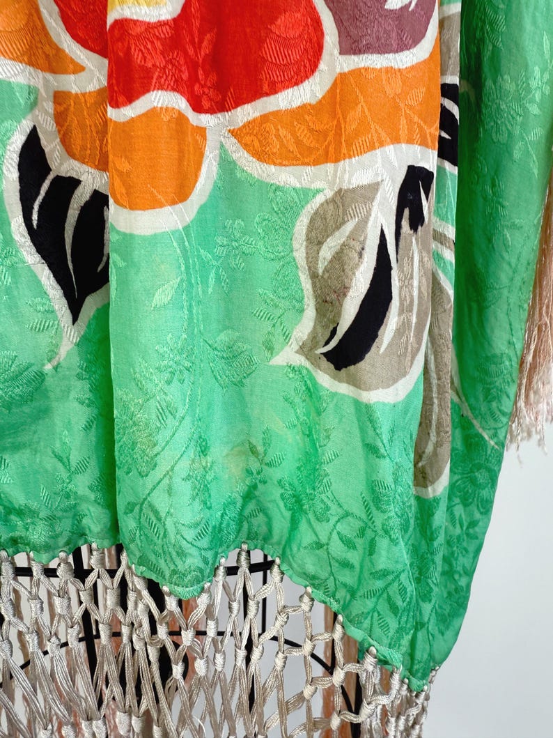 Puede incluir: Prenda vibrante con estampado floral en tonos verdes, naranjas, rojos y negros. La tela tiene un sutil dise&ntilde;o floral en relieve. El borde inferior presenta una borla color crema, que a&ntilde;ade textura y movimiento a la pieza.