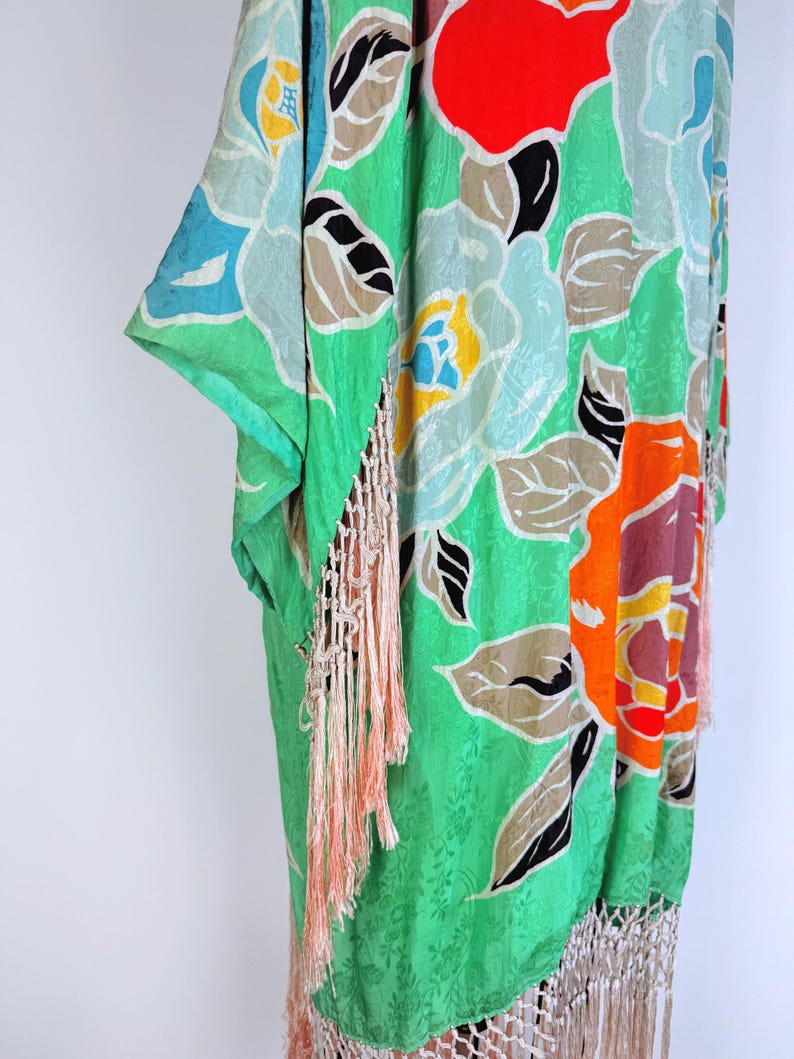 Puede incluir: Kimono floral verde con un vibrante estampado de flores rojas, azules y naranjas. El kimono presenta flecos melocot&oacute;n claro en el dobladillo y las mangas. La tela tiene un brillo sutil.