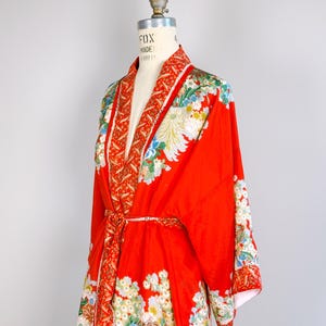 May include: A vibrant red kimono robe featuring a detailed floral pattern with white, blue, and green blossoms. The garment includes a wide collar with a contrasting orange and white leaf print, long sleeves, and a matching tie belt at the waist.