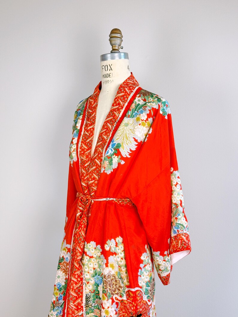 May include: A vibrant red kimono robe featuring a detailed floral pattern with white, blue, and green blossoms. The garment includes a wide collar with a contrasting orange and white leaf print, long sleeves, and a matching tie belt at the waist.