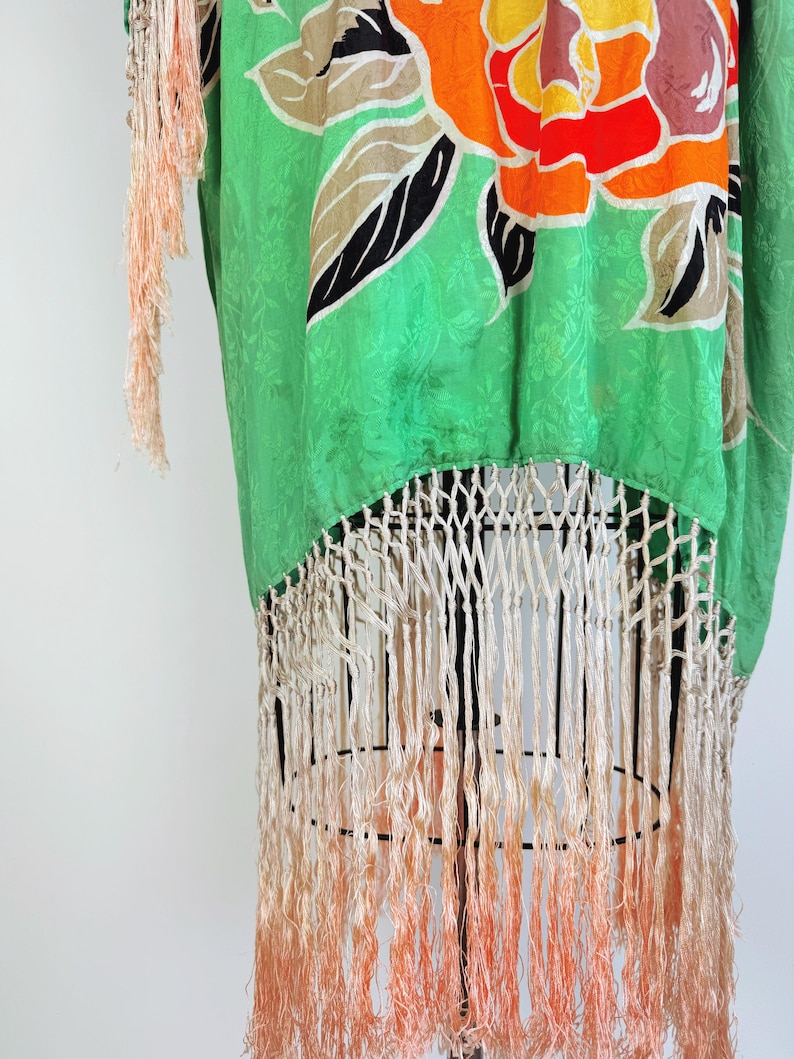 Puede incluir: Kimono de seda verde con un estampado floral en naranja, amarillo, marr&oacute;n y negro. El kimono tiene flecos color crema en la parte inferior con un efecto degradado, pasando de crema a melocot&oacute;n.