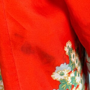 May include: A vibrant red fabric garment featuring a detailed floral pattern with white and light green blossoms. The textile displays a smooth texture with a visible dark stain on the upper left section of the red surface area.