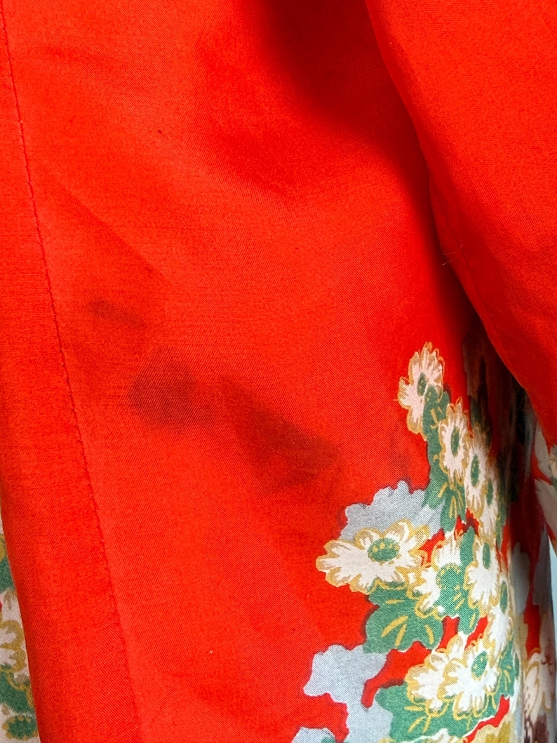 May include: A vibrant red fabric garment featuring a detailed floral pattern with white and light green blossoms. The textile displays a smooth texture with a visible dark stain on the upper left section of the red surface area.