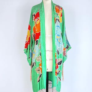Puede incluir: Una bata verde estilo kimono con un estampado floral en rojo, naranja, amarillo y azul. La bata tiene mangas largas y un dobladillo con flecos. La prenda se exhibe en un maniqu&iacute; sobre un fondo blanco.