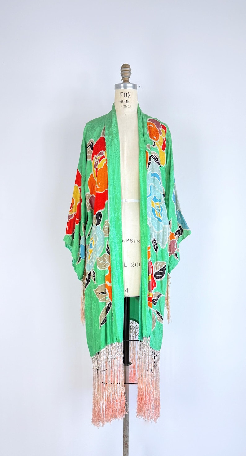 Puede incluir: Una bata verde estilo kimono con un estampado floral en rojo, naranja, amarillo y azul. La bata tiene mangas largas y un dobladillo con flecos. La prenda se exhibe en un maniqu&iacute; sobre un fondo blanco.