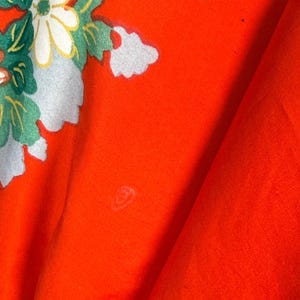 May include: A vibrant orange fabric featuring a printed floral pattern with white daisies, green leaves, and light blue accents. The textile displays a smooth, matte texture with visible folds and a small, faint circular mark on the surface.