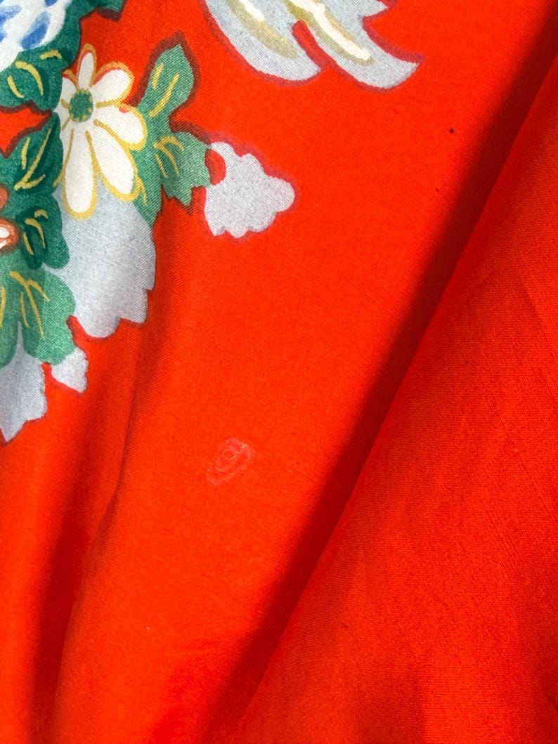 May include: A vibrant orange fabric featuring a printed floral pattern with white daisies, green leaves, and light blue accents. The textile displays a smooth, matte texture with visible folds and a small, faint circular mark on the surface.