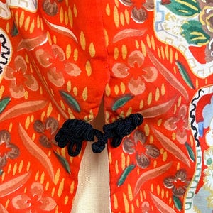 May include: A bright orange garment featuring a vibrant floral pattern in shades of yellow, green, and white. The piece is secured at the center with two black knotted fabric frog closures, creating a distinct contrast against the orange fabric.