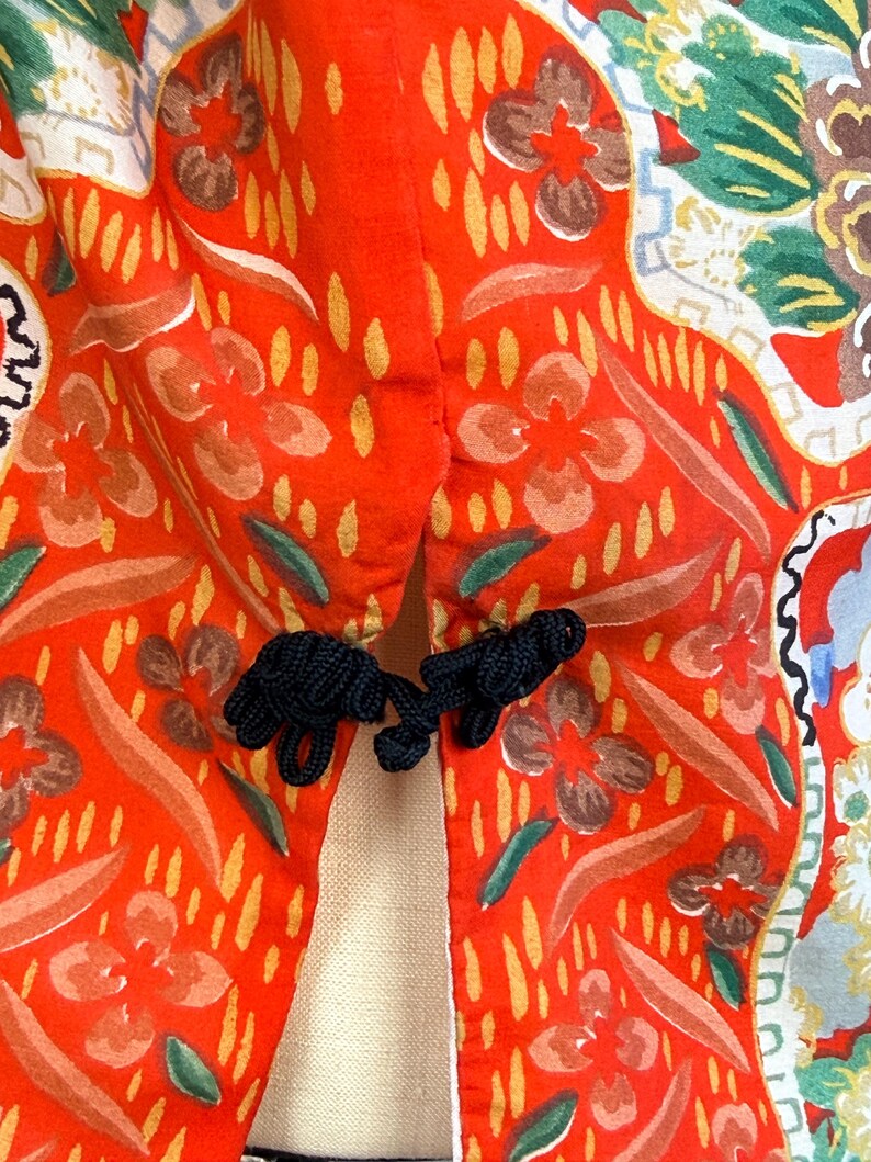 May include: A bright orange garment featuring a vibrant floral pattern in shades of yellow, green, and white. The piece is secured at the center with two black knotted fabric frog closures, creating a distinct contrast against the orange fabric.