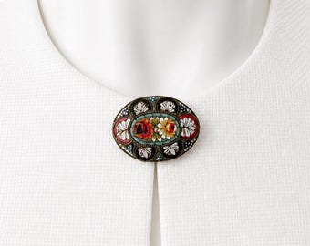 May include: Oval mosaic brooch with a floral design. The brooch features a central panel with red, yellow, and white flowers, surrounded by a border of white, red, and blue accents. The brooch is set against a white background.