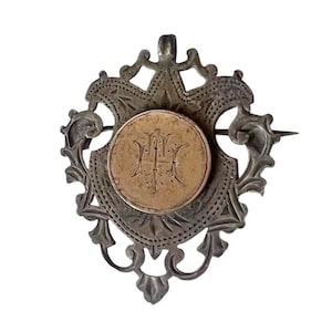 May include: Vintage silver brooch with a decorative shield-shaped frame and a central gold-coloured medallion. The medallion features an ornate monogram. The brooch has a pin closure and is likely antique jewellery.