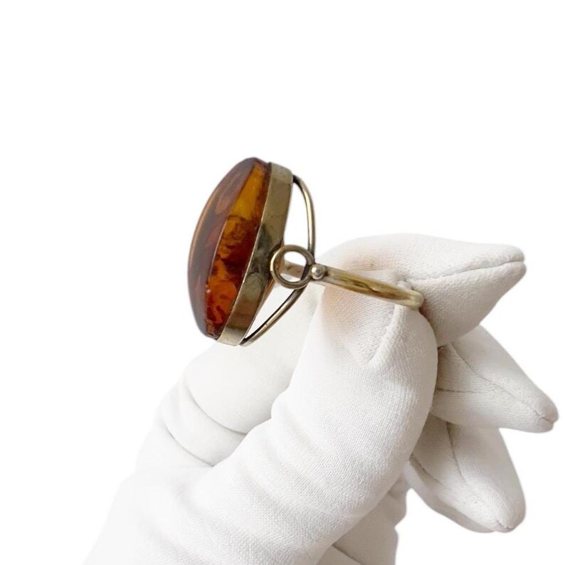 May include: A vintage ring featuring a large, oval amber stone set in a silver-toned metal frame. The amber has warm, golden-brown hues and is held by a simple, elegant band. The ring is shown against a white background.